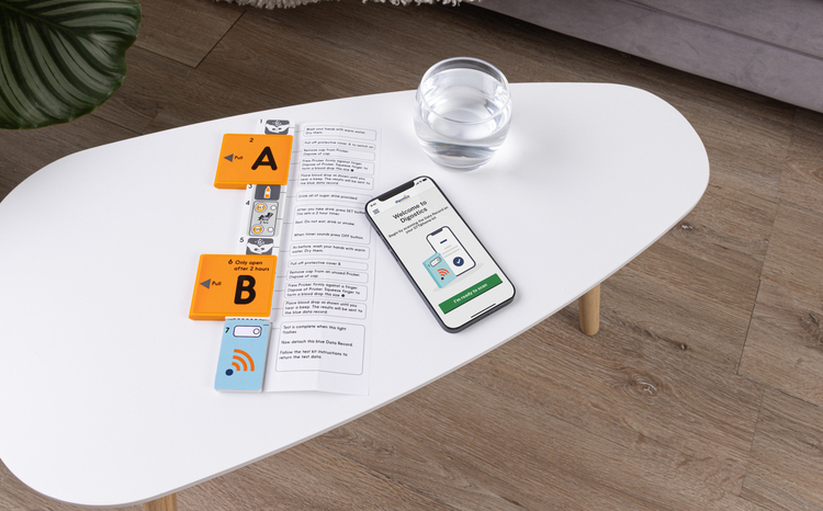 A GTT@home test kit, consisting of a smartphone app, glucose drink and electronic sensor device, laid out on an oval-shaped tabletop