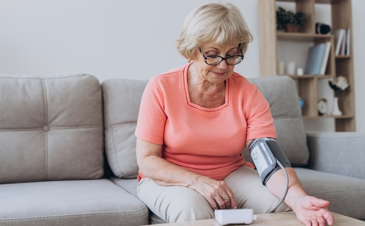 An older woman with glasses measuring her blood pressure in her living room using an arm pressure gauge and small medical device